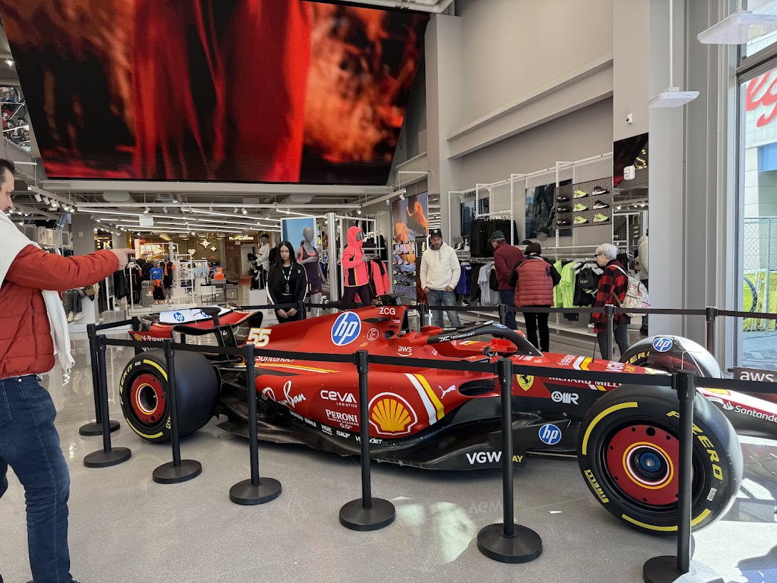 A open wheel red, black and yellow race car on display in a sports store with people admiring and point to it.