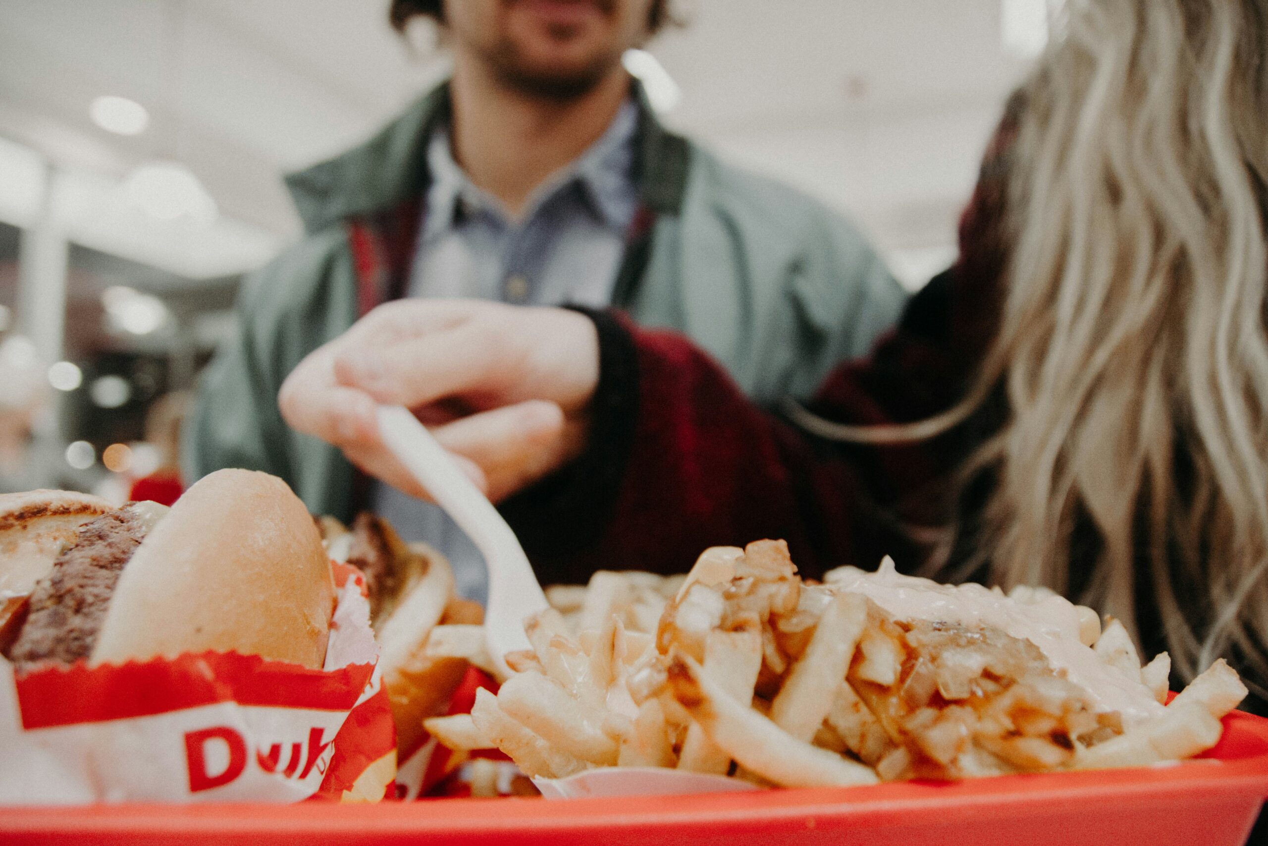 An image focused on a tray of fries and burgers with two people out of focus, eating the food